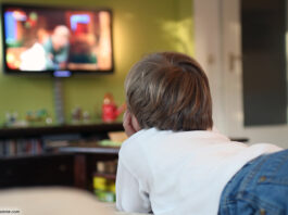 Kind liegt mit Fernbedienung in der Hand vor dem Fernseher