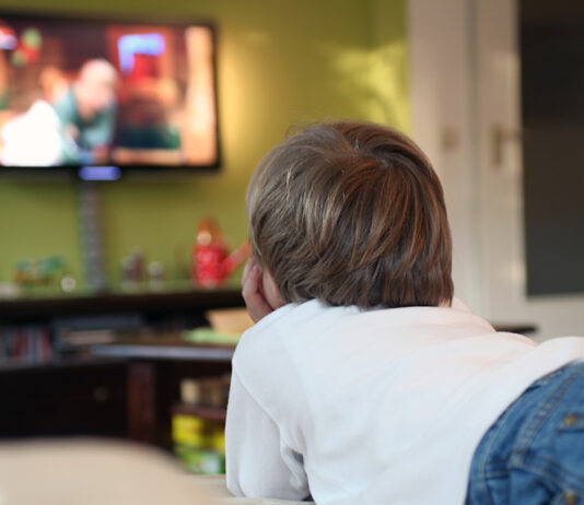 Kind liegt mit Fernbedienung in der Hand vor dem Fernseher