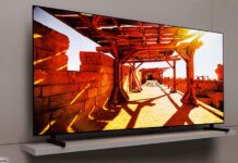 Ein 77 Zoll großer Samsung QD OLED TV vor einer beigen Wohnzimmerwand. Der TV-Bildschirm zeigt eine helle Spielfilmszene aus einem Außenbereich mit Schattenwürfen.