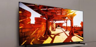 Ein 77 Zoll großer Samsung QD OLED TV vor einer beigen Wohnzimmerwand. Der TV-Bildschirm zeigt eine helle Spielfilmszene aus einem Außenbereich mit Schattenwürfen.