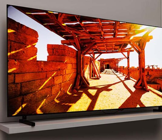 Ein 77 Zoll großer Samsung QD OLED TV vor einer beigen Wohnzimmerwand. Der TV-Bildschirm zeigt eine helle Spielfilmszene aus einem Außenbereich mit Schattenwürfen.