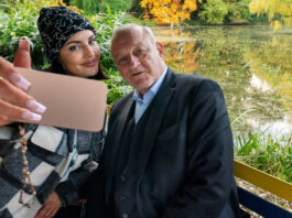 Wilsberg (Leonard Lansink) sitzt neben Bonnie von Laer (Johanna Ingelfinger) auf einer bunten Parkbank vor einem kleinen See. Bonnie holt ihr Handy vor sich und Wilsberg, um ein Selfie zu machen. Beide lächeln in die Handy-Kamera.