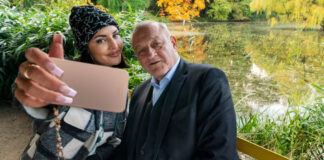 Wilsberg (Leonard Lansink) sitzt neben Bonnie von Laer (Johanna Ingelfinger) auf einer bunten Parkbank vor einem kleinen See. Bonnie holt ihr Handy vor sich und Wilsberg, um ein Selfie zu machen. Beide lächeln in die Handy-Kamera.