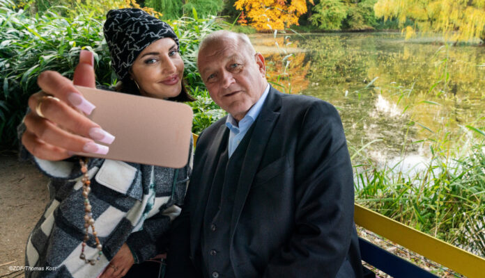 Wilsberg (Leonard Lansink) sitzt neben Bonnie von Laer (Johanna Ingelfinger) auf einer bunten Parkbank vor einem kleinen See. Bonnie holt ihr Handy vor sich und Wilsberg, um ein Selfie zu machen. Beide lächeln in die Handy-Kamera.