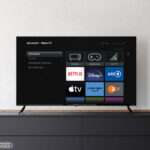 Ein Roku TV von Sharp in einem Beispielbild vor einer weißen Wand auf einem Sideboard