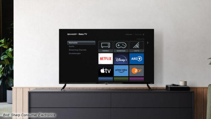 Ein Roku TV von Sharp in einem Beispielbild vor einer weißen Wand auf einem Sideboard