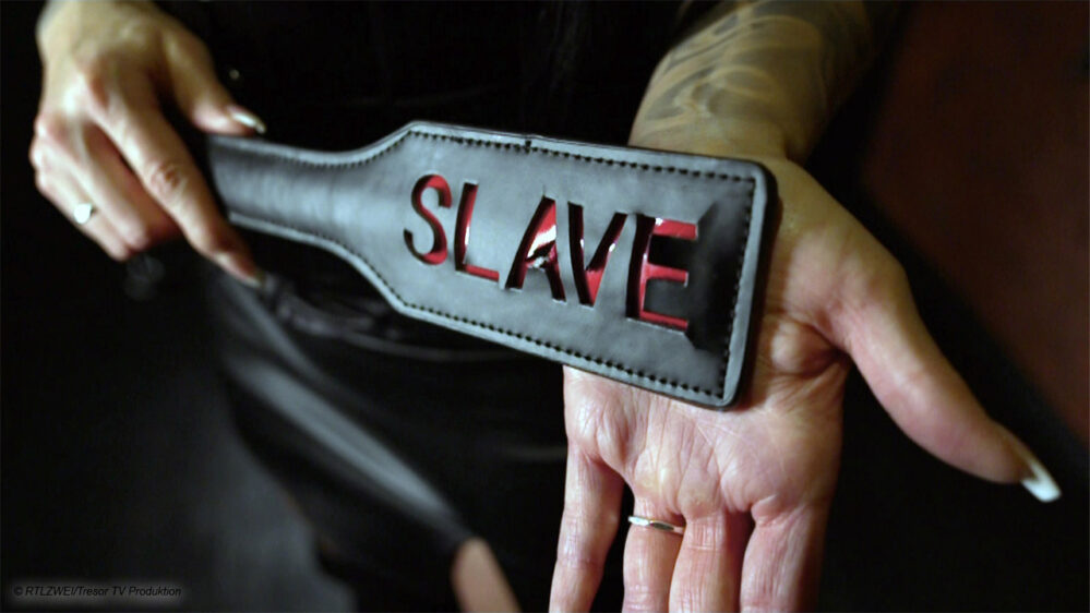 Lederpeitsche mit Gravur "Slave" vor einer Hand