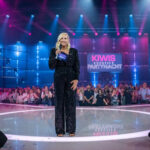 Andrea "Kiwi" Kiewel moderiert "Kiwis große Partynacht" bei Sat.1. Sie steht auf der Bühne vor dem Publikum und schaut in die Kamera.