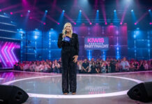 Andrea "Kiwi" Kiewel moderiert "Kiwis große Partynacht" bei Sat.1. Sie steht auf der Bühne vor dem Publikum und schaut in die Kamera.
