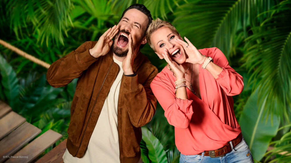 Dschungelcamp 2024: Jan Köppen und Sonja Zietlow stehen vor einer grünen Blätterwand und schreien belustigt in die Kamera.