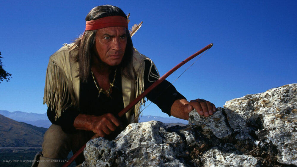 Winnetou steht mit einer hölzernen Waffe hinter einem Felsen.