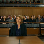 "Sie sagt. Er sagt.": Valerie Maiburg (Nicole Marischka) sitzt im Gerichtssaal im Zeugenstand und blickt zur Seite, hinter ihr sitzt das Publikum.