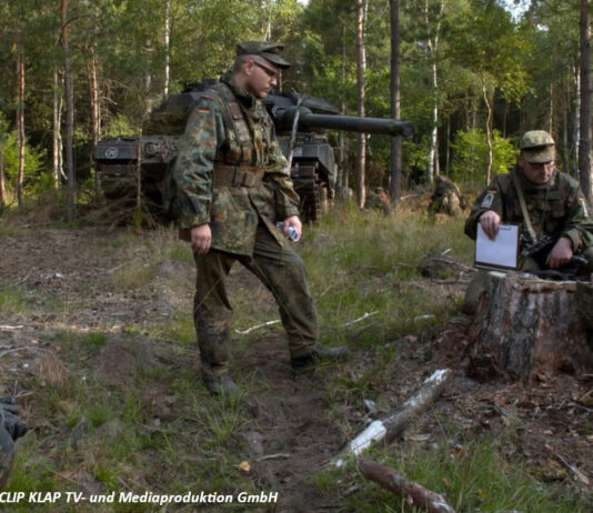 Soldaten sitzen bei einer Übung im Wald, im Hintergrund ein Leopard2-Panzer