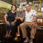 Susanne Daubner, Ina Müller und Götz Otto sitzen auf einem Tisch in einer Kneipe und schauen in die Kamera