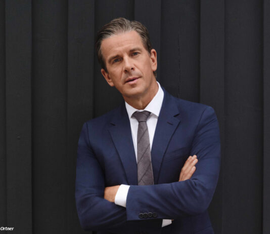 Moderator Markus Lanz steht mit verschränkten Armen vor einem anthrazitfarbenen Vorhang und schaut in die Kamera.