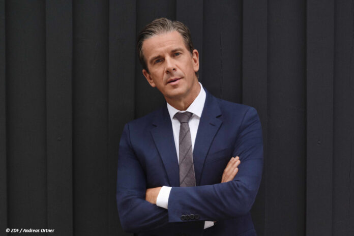 df_markus_lanz_2024 Moderator Markus Lanz steht mit verschränkten Armen vor einem anthrazitfarbenen Vorhang und schaut in die Kamera.