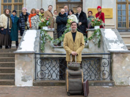 Sebastian Pastewka mit Koffer vor Schlosstreppe, auf der Hochzeitsgesellschaft steht. Aus Prime Video-Film "Die Brautentführung" (AT)