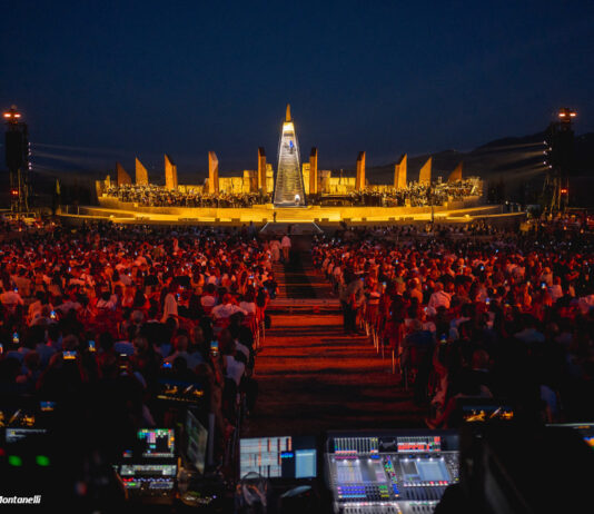 Eine Panorama-Aufnahme des Bühnenbilds des Jubiläum-Konzertes von Bocelli. Im Vordergrund steht das Kontrollpult für die Technik, dann kommen die bestzten Sitzplätze für die Zuschauenden und zentral dahinter das große, hell erleuchtete Bühnenbild.