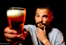 Ein Foto von exactly-Reporter Daniel Tautz vor schwarzem Hintergrund. Er hält ein volles Glas Bier in die Kamera und guckt skeptisch.