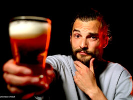 Ein Foto von exactly-Reporter Daniel Tautz vor schwarzem Hintergrund. Er hält ein volles Glas Bier in die Kamera und guckt skeptisch.