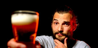 Ein Foto von exactly-Reporter Daniel Tautz vor schwarzem Hintergrund. Er hält ein volles Glas Bier in die Kamera und guckt skeptisch.