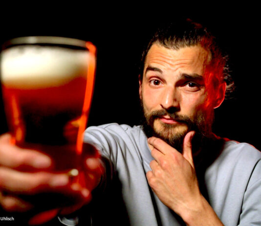 Ein Foto von exactly-Reporter Daniel Tautz vor schwarzem Hintergrund. Er hält ein volles Glas Bier in die Kamera und guckt skeptisch.