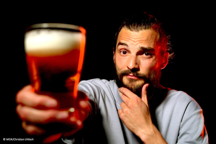 df_exactly_alltagsdroge_alkohol Ein Foto von exactly-Reporter Daniel Tautz vor schwarzem Hintergrund. Er hält ein volles Glas Bier in die Kamera und guckt skeptisch.