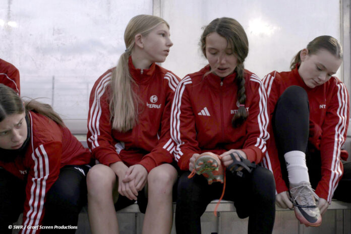 df_kick_it_like_women Vier Jugendliche in rot-weißen Trainingsanzügen sitzen auf einer Bank an einem Fußballplatz.