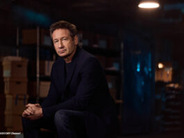 Der Schauspieler David Duchovny sitzt in einem abgedunkelten Raum und schaut in die Kamera.