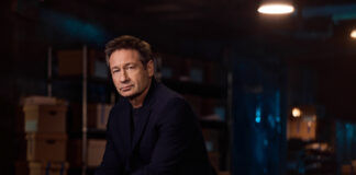 Der Schauspieler David Duchovny sitzt in einem abgedunkelten Raum und schaut in die Kamera.