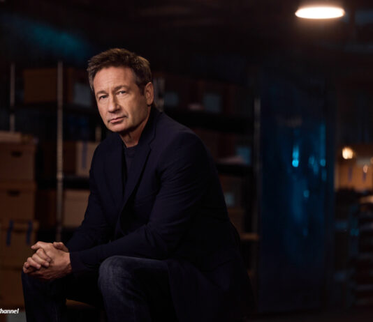 Der Schauspieler David Duchovny sitzt in einem abgedunkelten Raum und schaut in die Kamera.