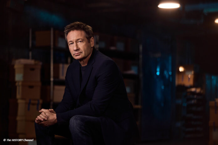 df_secrets_declassified_duchovny Der Schauspieler David Duchovny sitzt in einem abgedunkelten Raum und schaut in die Kamera.