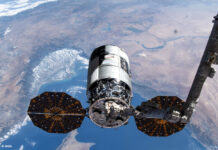Ein Cygnus-Raumfrachter wird von einem Metallarm im Weltraum gehalten. Im Hintergrund sieht man einen Teil der Erde.