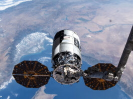 Ein Cygnus-Raumfrachter wird von einem Metallarm im Weltraum gehalten. Im Hintergrund sieht man einen Teil der Erde.