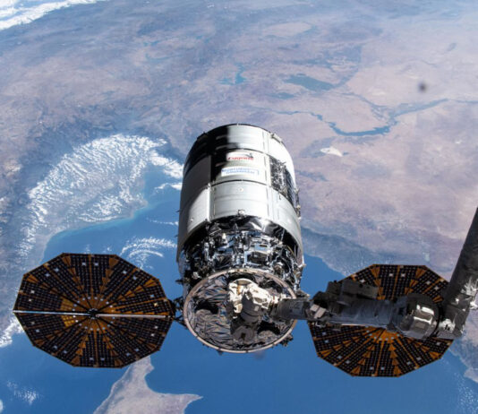 Ein Cygnus-Raumfrachter wird von einem Metallarm im Weltraum gehalten. Im Hintergrund sieht man einen Teil der Erde.
