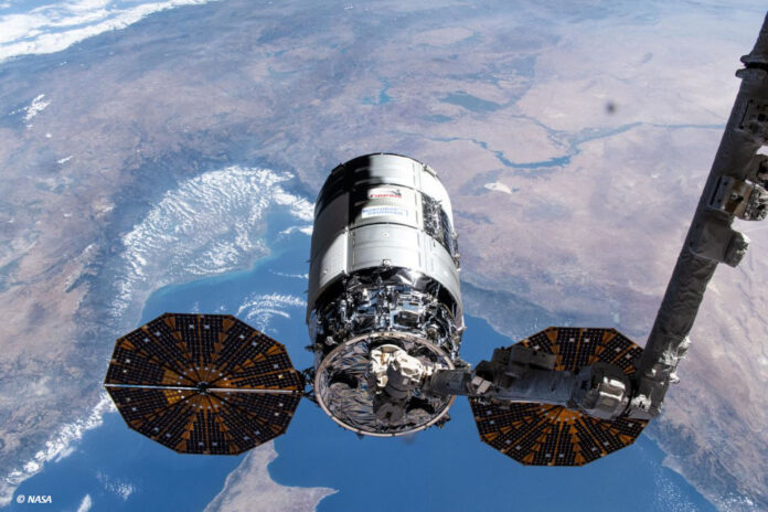 Ein Cygnus-Raumfrachter wird von einem Metallarm im Weltraum gehalten. Im Hintergrund sieht man einen Teil der Erde.